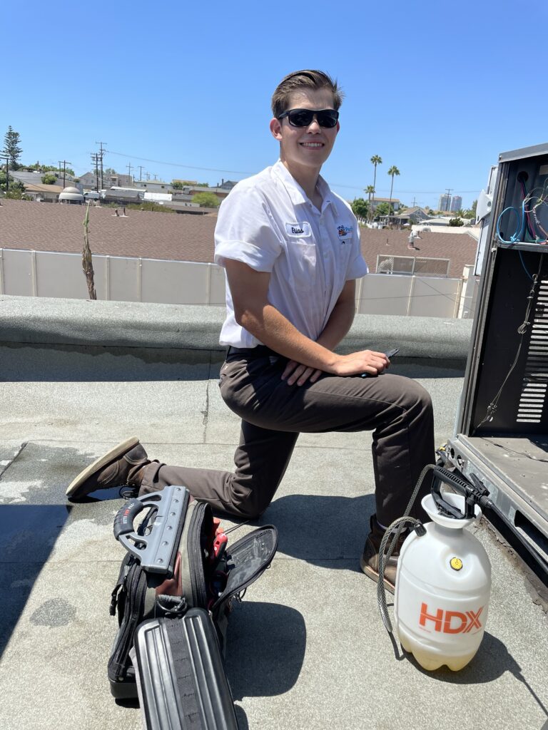 HVAC technician kneeling on a rooftop while servicing an air conditioning unit under a clear blue sky.